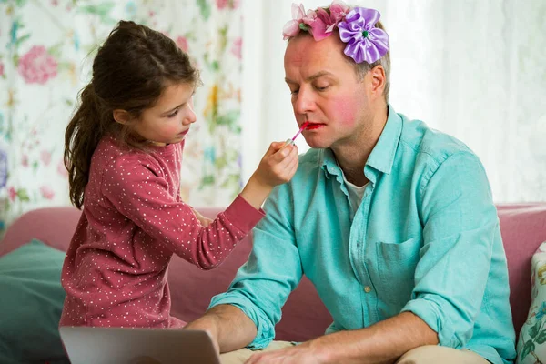 Child playing and disturbing father working remotely from home. Little girl applying makeup. Man sitting on couch with laptop. Family spending time together indoors.