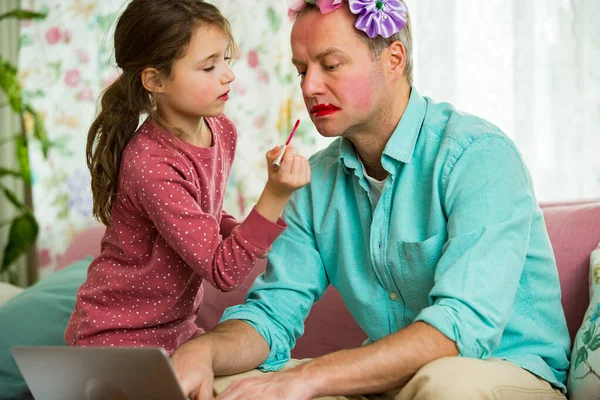 Child playing and disturbing father working remotely from home. Little girl applying makeup. Man sitting on couch with laptop. Family spending time together indoors.