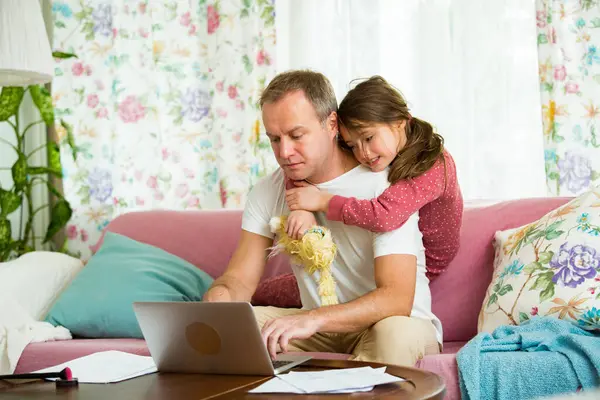 Child playing and disturbing father working remotely from home. Man sitting on couch with laptop. Family spending time together indoors.