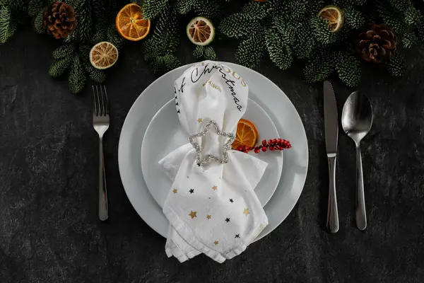 Christmas table setting, with white plates, silver cutlery, with a holiday napkin, on a dark relief background.