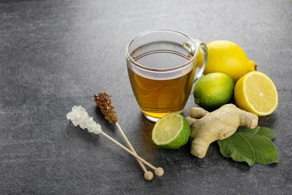 Herbal tea in a glass cup with sage, ginger, lemon and lime, brown sugar and sugar sticks against a dark background, close-up, side view. Copy space.