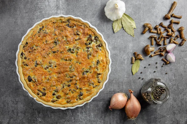 Traditional french pie Quiche Lorraine with mushrooms, potatoes, cheese, onions and seasonings. In a white baking dish, with forks, with black pepper and bay leaf, on a concrete background.Top view. — 스톡 사진