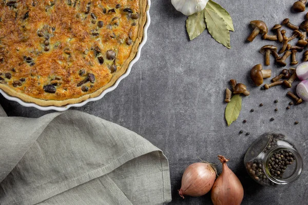 Traditional french pie Quiche Lorraine with mushrooms, potatoes, cheese, onions and seasonings. In a white baking dish, with forks, with black pepper and bay leaf, on a concrete background.Top view. — 스톡 사진