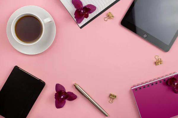 Feminine home office workspace. Tablet with copy space. Flat lay, social media, top view. Beauty business blog concept. Pink orchids flowers and tea, black coffee on pink background. Copy space.