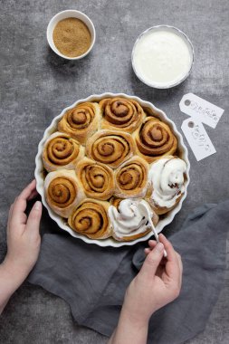 Tarçınlı çörek, kahverengi şekerli çörek, krema sosu ve tarçın çubukları. Kadın elleri çörekleri kremayla süsler. Üst Manzara. Gri beton arkaplan.