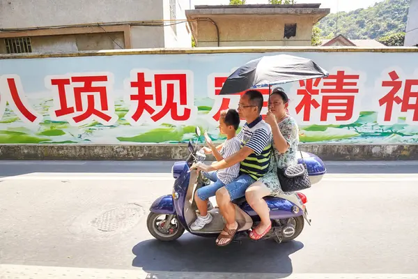 Three person family rides a scooter with umbrella protecting from the sun. — Stock Photo, Image