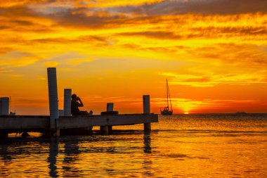 Genç adam Roatan 'ın günbatımını seyrediyor. Tahta bir rıhtımda oturuyor.