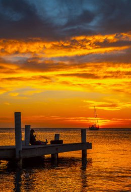 Genç adam Roatan 'ın günbatımını seyrediyor. Tahta bir rıhtımda oturuyor.