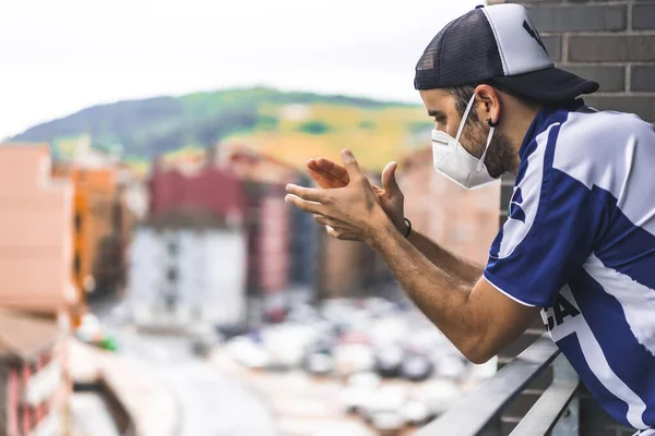 Maskeli ve mavi beyaz tişörtlü genç bir adam öğleden sonra saat 8 'de balkondaki tuvaletleri alkışlıyor. İspanya 'da pandemik koronavirüs