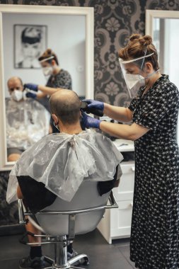 Kovid-19 için güvenlik önlemleri olan kuaför, yeni normallik, sosyal mesafe, maske takan kuaför ve müşteri. Makine saç kesiyor.