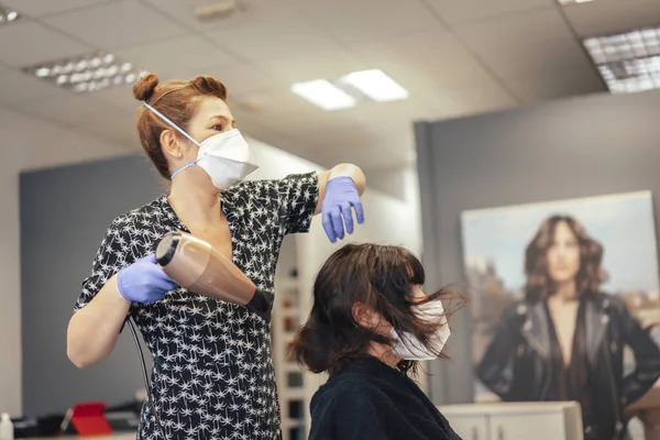 コヴィト 新しい正常性 社会的距離のためのセキュリティ対策を持つ美容師 マスクでブルネットのクライアントの髪を乾かす ロイヤリティフリーのストック写真