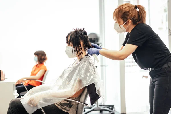 Hairdresser with mask and gloves working with a client and another in the background waiting. Reopening with security measures of Hairdressers in the Covid-19 pandemic