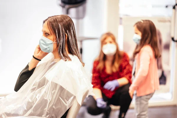 Hairdresser with mask and gloves talking to the client\'s daughter while the client answers an important call. Reopening with security measures for hairdressers in the Covid-19 pandemic