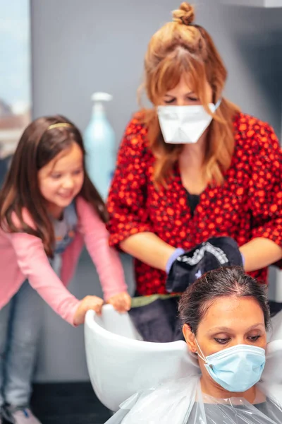 Hairdresser with mask and gloves washing the hair of the client and her daughter playing winged. Reopening with security measures for hairdressers in the Covid-19 pandemic