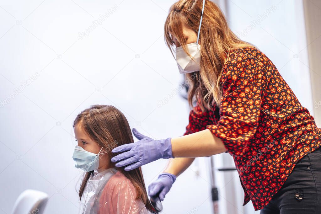 Hairdresser with mask and gloves combing the straight hair of a blonde girl, reflected in the mirror. Reopening with security measures for hairdressers in the Covid-19 pandemic