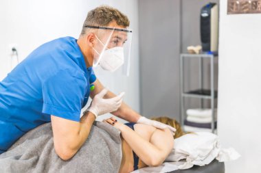 Ekran ve maskeli fizyoterapist genç bir kadına kalça masajı yapıyor. Covid-19 salgınında fizyoterapi güvenlik önlemleriyle tekrar açılıyor. Osteopati, tedavi edici kiromasaj.