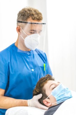 Maskeli ve ekranlı bir fizyoterapist maskeli bir çocuğa kafatası masajı yapıyor. Covid-19 salgınında fizyoterapi güvenlik önlemleri. Osteopati, tedavi edici kiromasaj.