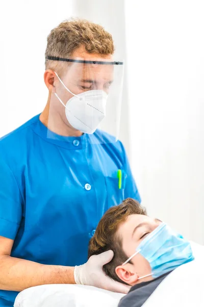 マスクとスクリーン付きの理学療法士は マスク付きの子供に頭蓋マッサージを与えます Covid 19パンデミックにおける理学療法の安全対策 変形性関節症 治療のカイロマッサージ — ストック写真