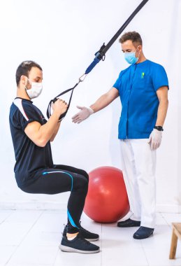 Mavi elbiseli bir fizyoterapist ve lastik bantlarla çalışan çömelmiş bir hasta. Kordemik Coronavirüs için koruyucu önlemler içeren fizyoterapi, spor masajı