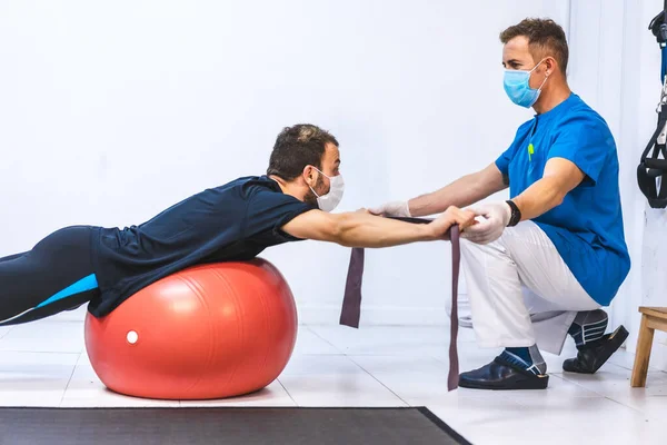 Maskeli bir fizyoterapist ve egzersiz yapan bir hasta. Koronavirüs salgını için koruyucu önlemlerle fizik tedavi, COVID-19. Osteopati, spor masajı