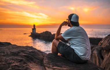 Beyaz gömlekli ve şapkalı genç bir adam San Sebastian, Gipuzkoa yakınlarındaki Pasajes San Juan deniz fenerinin turuncu gün batımına bakıyor. Bask Ülkesi