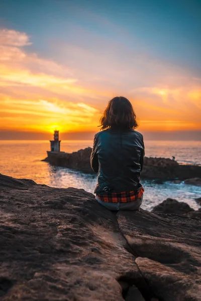 在圣胡安灯塔的橙色落日下 一位年轻女子穿着皮夹克坐在岩石上 — 图库照片