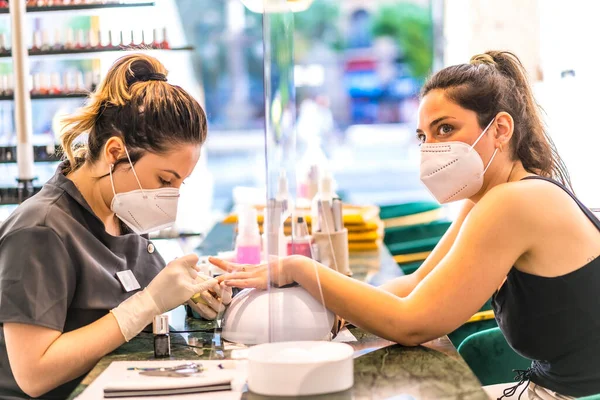 Yüzü maske takan güzellik salonu çalışanı esmer beyaz müşteriye renkli tırnaklar boyuyor. Korod-19 salgınından sonra tekrar açılıyor. Manikür ve Pedikür Salonu. Coronavirüs
