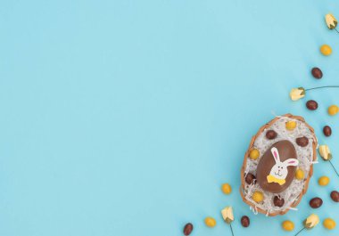 Yumurta sepetinde kahverengi ve sarı renkli paskalya yumurtaları ve mavi arka planda bir yuva ve bahar çiçeği gibi beyaz kağıtlar. Paskalya tavşanı büyük yumurtanın üstüne oturur. Mutlu Paskalyalar. Düzleştirme biçimi