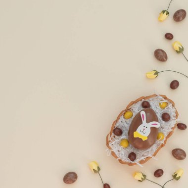 Yumurta sepetindeki renkli kahverengi ve sarı çikolatalı Paskalya yumurtaları, bej arka plandaki yuva ve bahar çiçekleri gibi beyaz kağıtlar. Paskalya tavşanı büyük yumurtanın üstüne oturur. Mutlu Paskalyalar. Düzleştirme biçimi