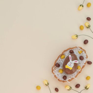 Yumurta sepetindeki renkli kahverengi ve sarı çikolatalı Paskalya yumurtaları, bej arka plandaki yuva ve bahar çiçekleri gibi beyaz kağıtlar. Paskalya tavşanı büyük yumurtanın üstüne oturur. Mutlu Paskalyalar. Düzleştirme biçimi