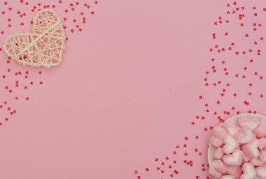 Sevgililer Günü konsepti. Rattan kalbi ve kalp şeklinde şekerlemeler pembe arka planda beyaz kasede, küçük kırmızı kalplerle. Sevgililer Günü tebrik kartı. Kopya alanı ile düz uzanma biçimi.