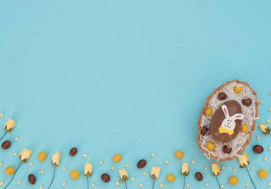 Yumurta sepetinde kahverengi ve sarı renkli paskalya yumurtaları ve mavi arka planda bir yuva ve bahar çiçeği gibi beyaz kağıtlar. Paskalya tavşanı büyük yumurtanın üstüne oturur. Mutlu Paskalyalar. Düzleştirme biçimi