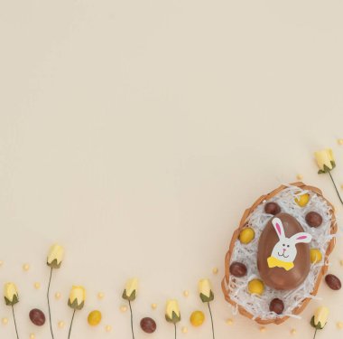 Yumurta sepetindeki renkli kahverengi ve sarı çikolatalı Paskalya yumurtaları, bej arka plandaki yuva ve bahar çiçekleri gibi beyaz kağıtlar. Paskalya tavşanı büyük yumurtanın üstüne oturur. Mutlu Paskalyalar. Düzleştirme biçimi