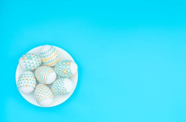 Süslü Paskalya yumurtaları mavi arka plandaki beyaz tabakta yatar. Mutlu Paskalya tatili konsepti. Selamlar, davetiye. Kopya alanı ile düz uzanma biçimi.