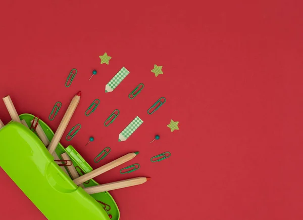 Green pencil case, wooden pencils, red and green clips and pins, pencil and star shaped paper stickers on red background. School and office supplies, stationary. Back to school concept. Flat lay style