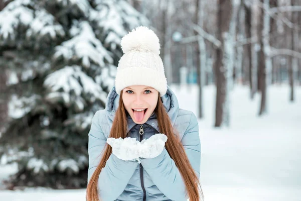 Una chica vestida con ropa abrigada de invierno y un sombrero posando en un bosque de invierno. Retrato de una joven mostrando su lengua . — Foto de Stock