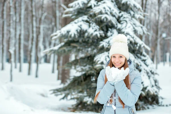 一个女孩穿着暖和的冬天的衣服和帽子在冬天的森林里吹雪, 水平。模型与美丽的微笑在圣诞树附近. — 图库照片