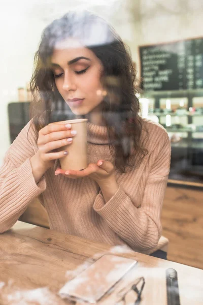 Chica sostiene una taza de papel con café sentado en una mesa de madera en una cafetería. Chica con una taza de café en las manos, cerró los ojos y disfruta del aroma, detrás del vaso en un café . — Foto de Stock