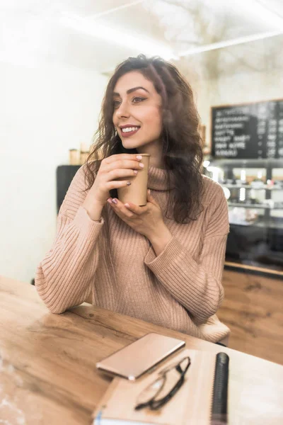 Chica sostiene una taza de papel con café sentado en una mesa de madera en una cafetería — Foto de Stock