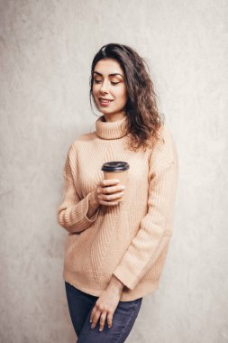 Sıcak süveter giymiş, elinde bir fincan kahveyle duvarın önünde duran genç ve güzel bir kız. Esmer ve bir fincan kahve..