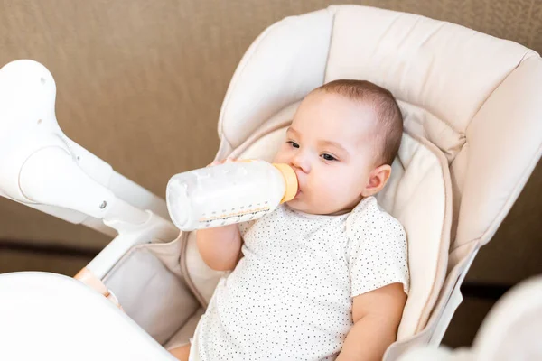Baby eats in a highchair. Mom feeds the baby with pureed food. The first feeding of the baby. The kid eats and drinks from the bottle on his own.