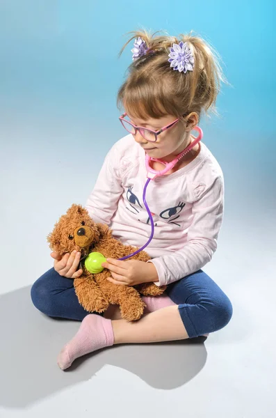 Chica bonita juega en el médico trata a un oso de peluche en una ba gris — Foto de Stock