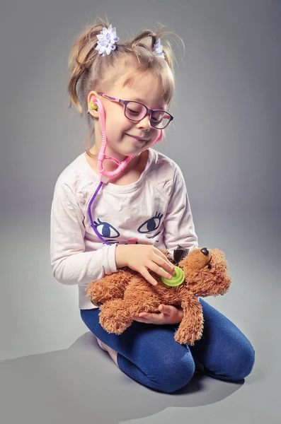 Chica bonita juega en el médico trata a un oso de peluche en una ba gris — Foto de Stock