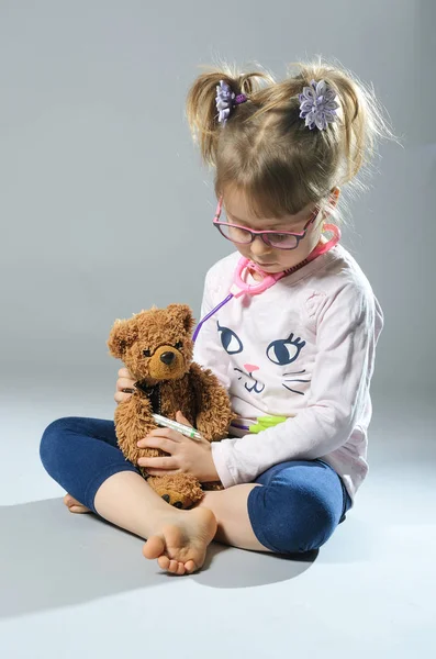 Chica bonita juega en el médico trata a un oso de peluche en una ba gris — Foto de Stock