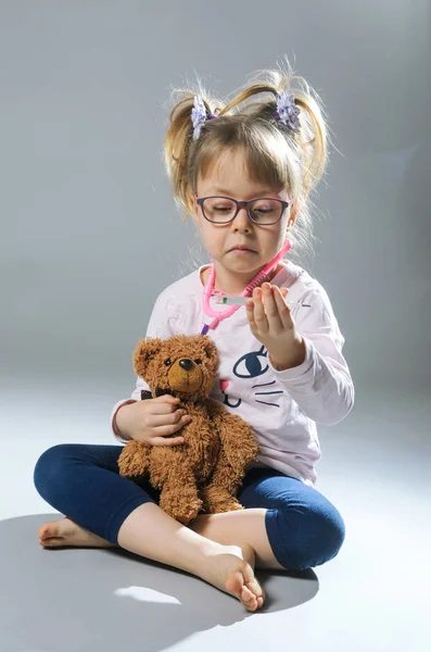 Chica bonita juega en el médico trata a un oso de peluche en una ba gris — Foto de Stock