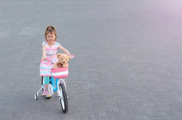 Una hermosa niña está montando una bicicleta con un oso de peluche en — Foto de Stock