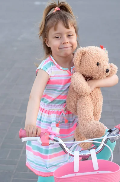 एक सुंदर छोटी लड़की एक टेडी भालू के साथ साइकिल पर सवारी कर रही है . — स्टॉक फ़ोटो, इमेज