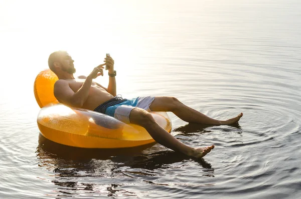 Un hombre sentado en un anillo inflable en el agua y sosteniendo un sm — Foto de Stock