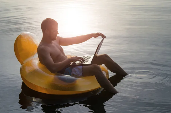 Hombre con portátil en un anillo de goma en el agua al atardecer, sp gratis — Foto de Stock