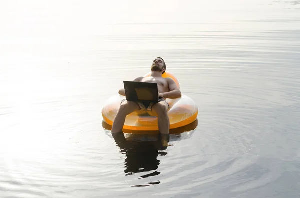En man med en laptop på en uppblåsbar ring i vattnet vid solnedgången — Stockfoto